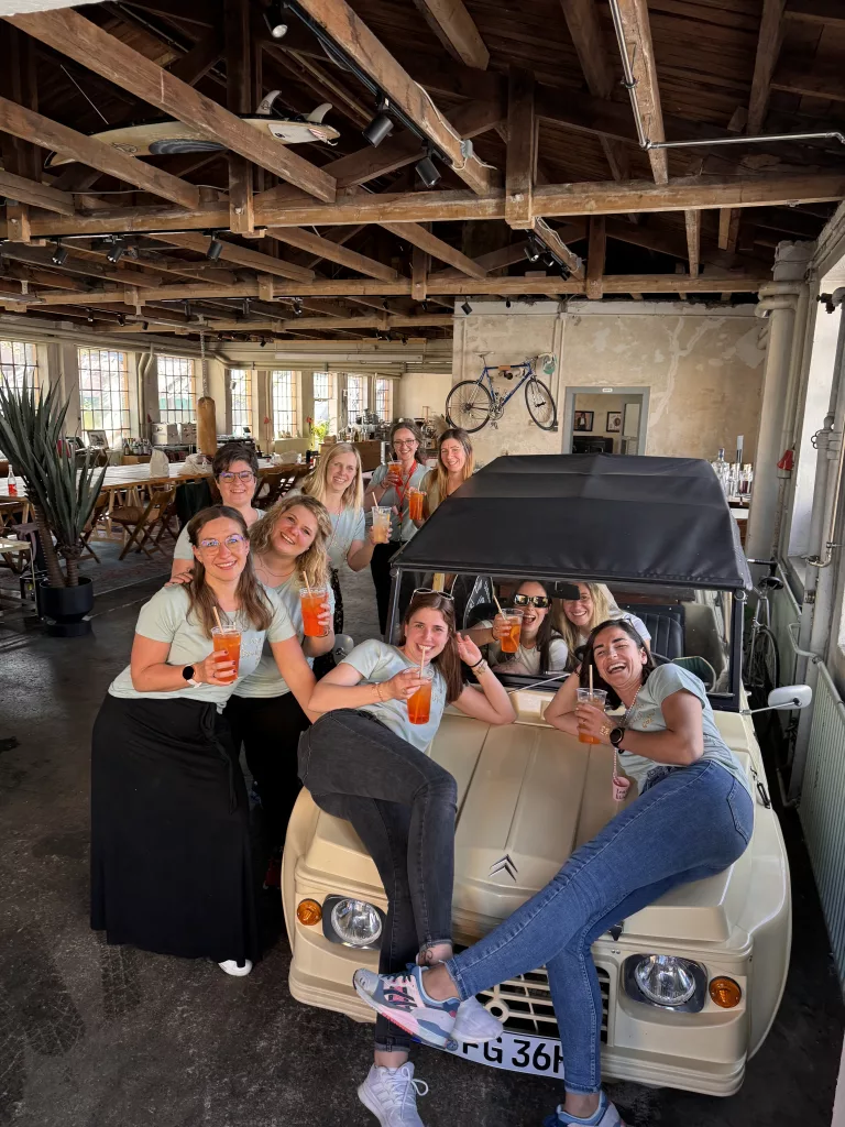 Ausgelassene Gruppe von Frauen beim JGA, die mit Aperol Spritz auf einem Renault Mehari in einem Loft mit Holzbalken posieren.